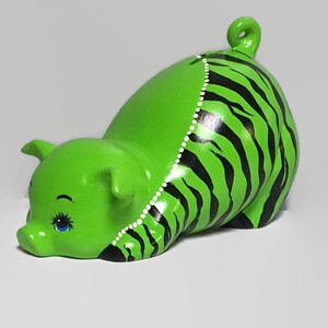 May include: Green ceramic piggy bank with black and white stripes. The piggy bank has a curled tail and one blue eye.