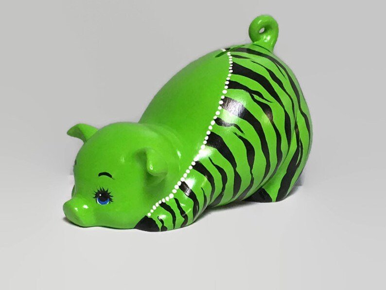 May include: Green ceramic piggy bank with black and white stripes. The piggy bank has a curled tail and one blue eye.
