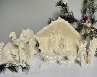 May include: A white ceramic nativity scene with a stable, three wise men, a camel, a donkey, sheep, and angels. The figures are all in a white, pearlescent finish.