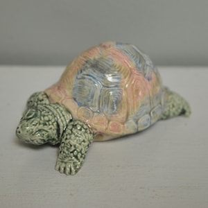 May include: A ceramic turtle figurine with a colorful, multi-colored shell. The shell is painted in shades of blue, pink, and yellow. The turtle's head and legs are green.
