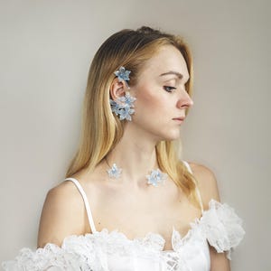 May include: A woman wearing a white corset top with lace trim and a matching skirt. She is also wearing a set of light blue flower-shaped jewelry, including earrings and a necklace. The background is a solid, neutral color.
