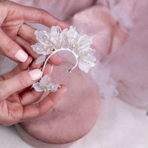 Può includere: Un delicato orecchino a cerchio floreale e un orecchino abbinato. I petali bianchi traslucidi sono accentuati da filo argentato e cristalli scintillanti. I gioielli sono tenuti su uno sfondo rosa tenue, suggerendo un tema nuziale o per occasioni speciali.