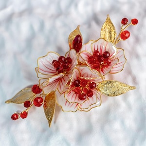 May include: A floral hair accessory featuring white and red flowers with gold wire outlines. The design includes gold-colored leaves and clusters of red beads, creating an elegant and decorative piece.