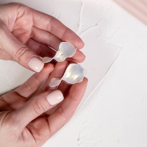 Puede incluir: Un par de pendientes iridiscentes en forma de pétalo con un acabado translúcido y nacarado. Los pendientes presentan una forma curva y orgánica que imita la apariencia de delicados pétalos de flor, captando la luz con sutiles cambios de color.