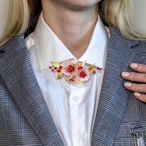 May include: A floral brooch featuring pink and white flowers with red accents and gold-colored leaves. The brooch is pinned to a white collared shirt, worn under a gray plaid blazer.