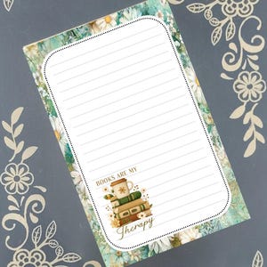 May include: A rectangular notepad featuring a floral pattern with white daisies and green foliage. The center has a white area with horizontal lines for writing. At the bottom, there is an illustration of stacked books, a mug, and the text "BOOKS ARE MY Therapy."