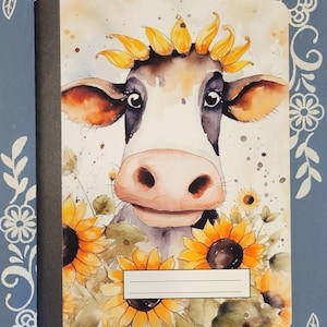 Puede incluir: Una ilustración de acuarela de una vaca con una corona de girasoles. La vaca está rodeada de girasoles y vegetación. La imagen está en una cubierta de cuaderno.