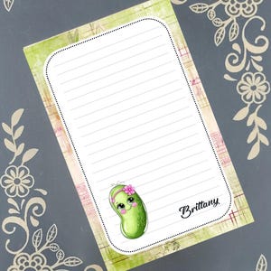 May include: A lined notepad featuring a cartoon pickle character with a pink flower headband. The notepad has a green and pink border, with the name "Brittany" in cursive. Floral decorative elements frame the notepad.