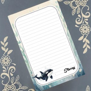 May include: A lined notepad with a watercolor design featuring an orca whale. The notepad has a dotted border and the name "Stacey" in script. The background has a blue and green watercolor effect.