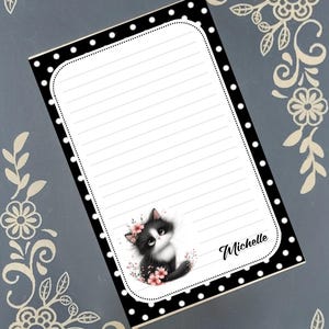 May include: Black and white notepad with a black and white polka dot border. The notepad has a black and white cat illustration with pink flowers. The notepad has lined paper and the name "Michelle" written at the bottom.