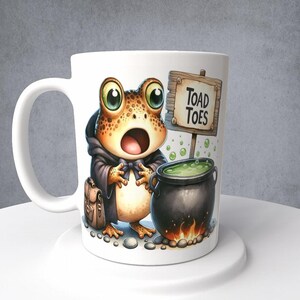 May include: White ceramic mug with a cartoon illustration of a frog wearing a black robe and holding a cauldron. The frog has a surprised expression and the text "Toad Toes" is on a sign above the cauldron.