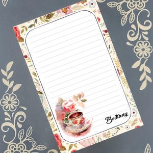 May include: A lined notepad with a floral border and a watercolor illustration of a teacup and roses. The name "Brittany" is written in cursive. The notepad is on a gray background with floral corner designs.