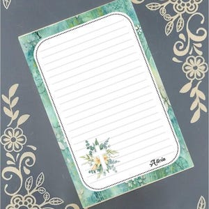 Può includere: Un blocco note verde e bianco con righe, con un design floreale e una croce bianca. Il nome "Alicia" è scritto in fondo al blocco note.