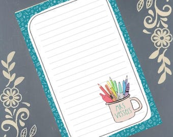 May include: A lined notepad with a teal border featuring a pattern of small shapes. A mug filled with colorful pencils is in the lower right corner, with the text "MRS Vessells" on the mug. The notepad is on a gray background with floral corner designs.