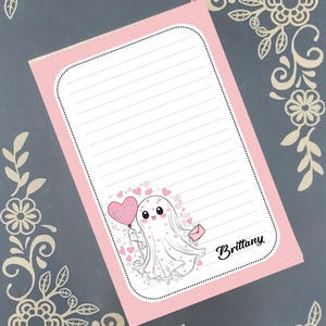May include: A pink lined notepad featuring a cartoon ghost holding a heart-shaped balloon with the text "Be My Boo". The notepad also has a small pink envelope and the name "Brittany". The design is decorated with hearts.