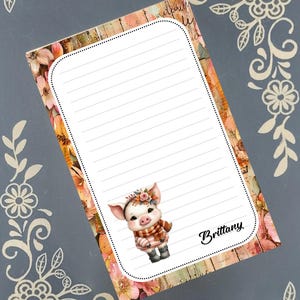 May include: A lined notepad with a floral and wood-grain border. A cute piglet wearing a scarf and flower crown is featured on the notepad. The name "Brittany" is written in cursive on the notepad.