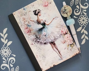 Könnte beinhalten: Ein Notizbuch mit einer Ballerina-Illustration auf dem Cover. Die Ballerina trägt ein hellblaues und weißes Tutu und ist von Blumen umgeben. Das Notizbuch liegt auf einer blauen Oberfläche mit einem weißen Stift und einem dekorativen Anhänger.
