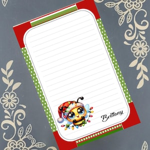 May include: A lined notepad with a red and green border, featuring a cartoon bee wearing a Santa hat and Christmas lights. The notepad has the name "Brittany" in script.