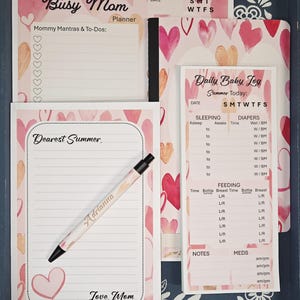 May include: A pink and white planner set for a busy mom, featuring a planner, notepad, and baby log. The set includes a pen with the name "Adrianna" and is decorated with hearts. The planner includes sections for daily tasks and baby care.