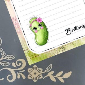 May include: A notepad featuring a cartoon pickle character with a pink flower crown. The notepad has lined paper and the name "Brittany" in cursive. The background is a grey surface with floral designs.