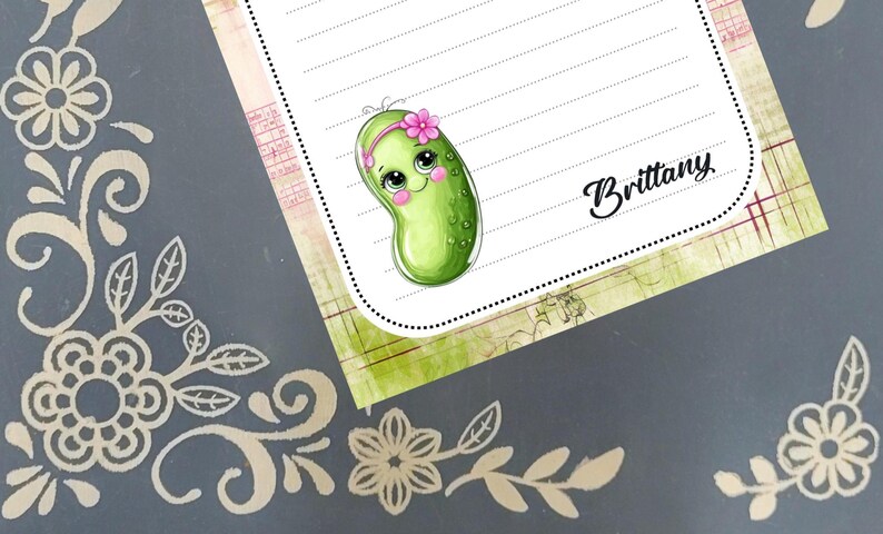 May include: A notepad featuring a cartoon pickle character with a pink flower crown. The notepad has lined paper and the name "Brittany" in cursive. The background is a grey surface with floral designs.