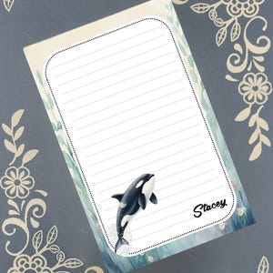 May include: Lined notepad featuring a black and white orca leaping from the water. The notepad has a watercolor background with a blue and green color scheme. The name "Stacey" is written in cursive. Decorative floral elements frame the notepad.