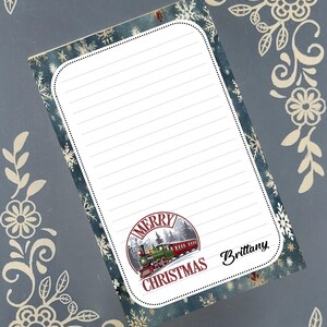 Può includere: Blocco note rettangolare a tema natalizio. Presenta un'illustrazione di un treno con la scritta "Merry Christmas". Lo sfondo ha un motivo a fiocchi di neve. Il blocco note è a righe per la scrittura.