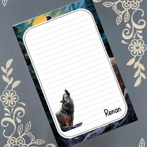 May include: Lined notepad featuring a howling wolf illustration. The notepad has a white border with a dotted design and the name "Ronan" in the bottom right corner. The background shows a mountain scene with a yellow and blue sky.