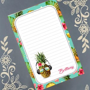 May include: A lined notepad with a turquoise background featuring pineapples, tropical leaves, and pink hibiscus flowers. A cartoon pineapple wearing sunglasses and headphones is on the notepad. The name "Brittany" is written in pink script.