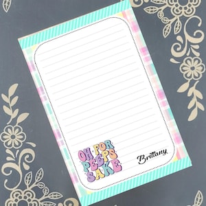 May include: A lined notepad with a pastel plaid and striped border. The notepad features the text "OH FOR PEEPS SAKE" in colorful bubble letters and the name "Brittany" in black script. The background is a dark gray with floral corner designs.