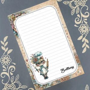 May include: A lined notepad with a floral and musical note background. The notepad features a cartoon cat chef holding a rolling pin and knife. The name "Brittany" is written in cursive on the notepad.