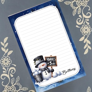 May include: A lined notepad with a blue watercolor border and a snowman illustration. The snowman wears a top hat and scarf, holding a bucket of snowballs. A sign reads "Snowballs 5 Cents". The name "Brittany" is printed on the notepad.