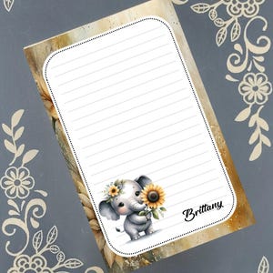 Könnte beinhalten: Ein liniertes Notizblock mit einer Illustration eines Babyelefanten, der eine Sonnenblume hält. Der Elefant trägt einen Blumenkranz. Der Notizblock hat den Namen "Brittany". Der Hintergrund ist eine Mischung aus Gold und Braun.
