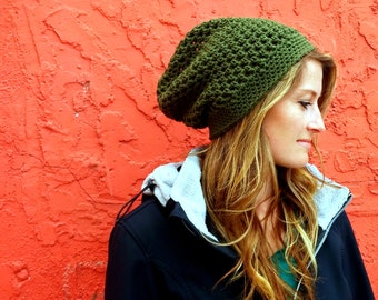 May include: A green crocheted beanie hat with a slouchy fit. The hat is worn by a person with long blonde hair and a black jacket.