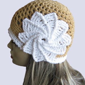Könnte beinhalten: Ein gehäkelter Hut in Beige mit einem weißen Blumenmuster. Der Hut hat einen weißen Rand und eine große, spiralförmige weiße Blume an der Seite. Der Hut ist handgefertigt und hat ein strukturiertes Aussehen.