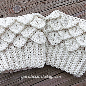 May include: Two off-white, crocheted wrist cuffs. The cuffs have a ribbed base and a scalloped, scale-like pattern along the top edge. The cuffs are handmade and appear soft and textured. The background is a wooden surface.
