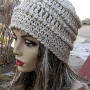 Könnte beinhalten: Beige Häkelmütze mit geripptem Rand. Die Mütze ist aus dickem, strukturiertem Garn gefertigt und hat eine lockere Passform. Die Mütze wird auf einer Schaufensterpuppe mit langen, hellbraunen Haaren gezeigt.