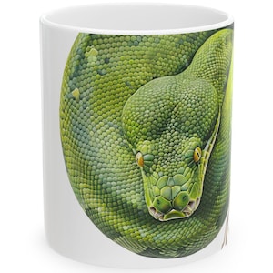 Könnte beinhalten: Weiße Keramiktasse mit einer detaillierten Illustration einer aufgerollten grünen Baumpython. Die Schuppen und gelben Augen der Schlange sind lebendig dargestellt. Die Tasse hat eine Standardgröße, ideal für Kaffee oder Tee.