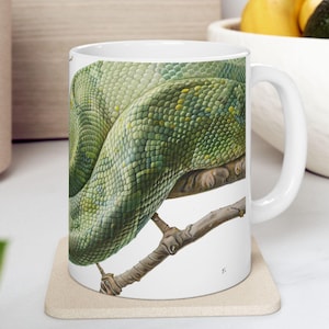 May include: White ceramic mug featuring a detailed illustration of a green snake coiled around a branch. The snake's scales are a mix of green and yellow. The mug sits on a beige coaster.