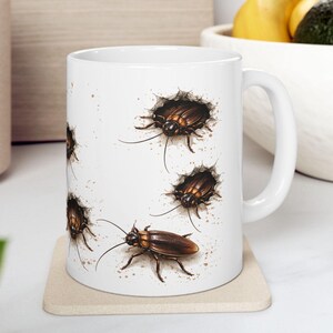 May include: White ceramic mug featuring a detailed illustration of several brown cockroaches. The insects are depicted emerging from holes, with a realistic, artistic style. The mug is on a beige coaster, with a white handle.