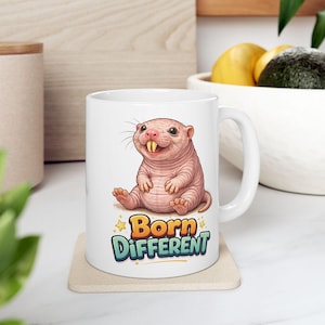 Op de afbeelding: Witte keramische mok met een cartoon naakte molrat en de tekst "Born Different". De mok staat op een beige onderzetter. Het ontwerp toont een roze molrat met grote tanden.