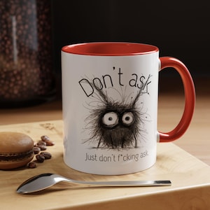 Puede incluir: Taza de cerámica blanca con interior y asa rojos. La taza presenta una ilustración de monstruo de dibujos animados con el texto "Don't ask. Just don't f*cking ask." Una cuchara y un pastelito están en primer plano.