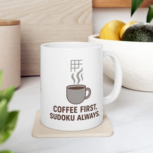 May include: White ceramic mug with a gray coffee cup graphic and the text "COFFEE FIRST. SUDOKU ALWAYS." The mug also features a gray sudoku grid and steam graphic. The mug is on a beige coaster.