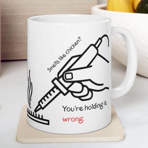 Pode incluir: Caneca de cerâmica branca com um gráfico preto e vermelho. O gráfico mostra uma mão segurando uma seringa sobre uma grelha com o texto "Smells like chicken? You're holding it wrong."