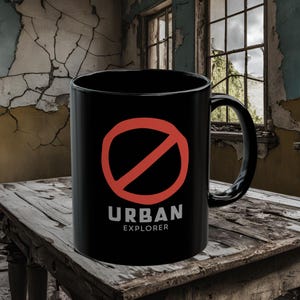 Könnte beinhalten: Schwarze Kaffeetasse mit einem roten Kreis und einem diagonalen Strich durch ihn. Der Text "URBAN EXPLORER" ist unter dem Kreis gedruckt.