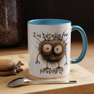 Puede incluir: Taza de cerámica blanca con interior y asa azules. La taza presenta una ilustración de dibujos animados de una criatura espinosa con ojos grandes y el texto "I'm totally fine... probably."