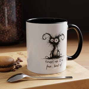 May include: White ceramic mug with a black interior and handle. The mug features a whimsical black and white illustration of a creature with large eyes and the text "Trust me. I'm fine... kind of."