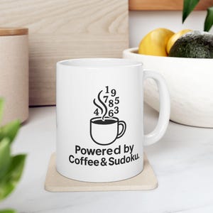 May include: White ceramic mug with a black graphic of a coffee cup, steam, and numbers. The text "Powered by Coffee & Sudoku" is below the cup. The mug sits on a beige coaster.