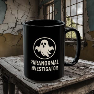 Könnte beinhalten: Schwarze Kaffeetasse mit einem weißen Geistermotiv und dem Text "Paranormal Investigator".