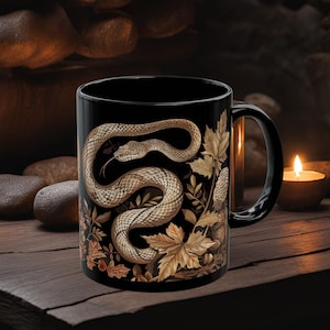 Puede incluir: Taza de café negra con una ilustración detallada de una serpiente enroscada alrededor de hojas de otoño y bellotas. La serpiente es marrón y blanca sobre un fondo negro.